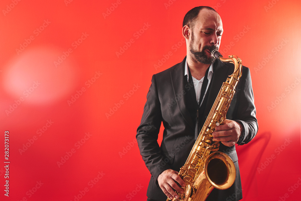 Naklejka premium Portrait of professional musician saxophonist man in suit plays jazz music on saxophone, red background in a photo studio