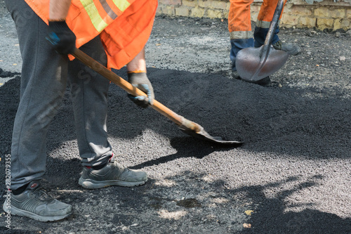 Worker man lays asphalt road repair road paving yellow sun ray light. A man in overalls is laying asphalt with a shovel