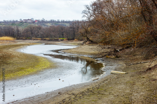 The dry river bed with small remains of water with leafless forest on the shore