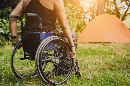 Disabled man resting in a campsite with friends. Wheelchair in the forest on the background of tents