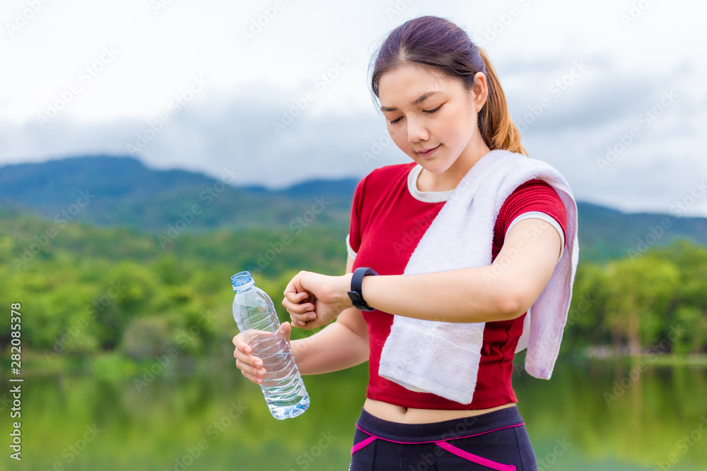 Beautiful young Asian woman looking at her smart watch and while holding a water bottle to drink after her exercise run at a local lake, healthy lifestyle Beautiful young Asian woman looking at her smart watch and while holding a water bottle to drink after her exercise run at a local lake, healthy lifestyle
