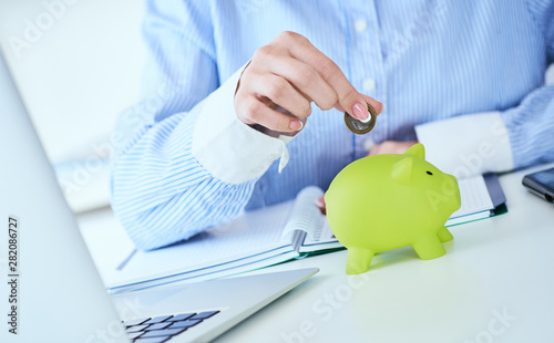 Woman's hand putting money coin in green piggy bank close-up in office background. Growing business, pension and insurance savings concept.