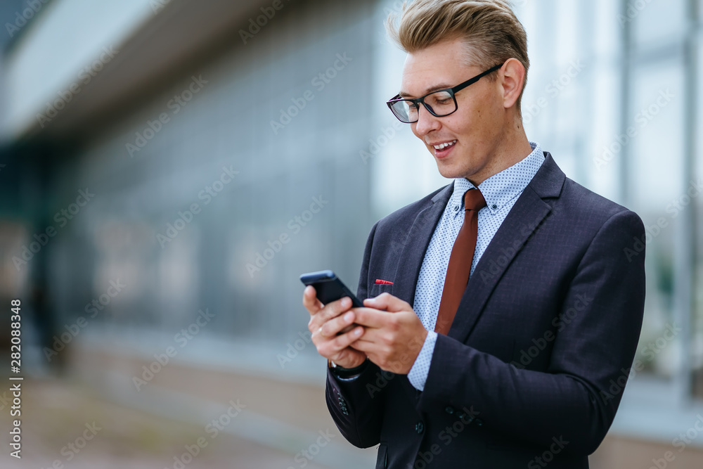 Happy businessman smiling as he reads a text message standing outside the office. Male executive manager in trendy clothes using smartphone outdoors. Modern technology concept Happy businessman smiling as he reads a text message standing outside the office. Male executive manager in trendy clothes using smartphone outdoors. Modern technology concept