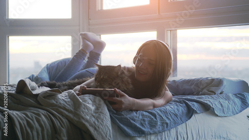 Young woman in headphones relaxing with her lovely big Maine Coon cat makes selfie laying down in bed by the window during sunset with lens flare effects