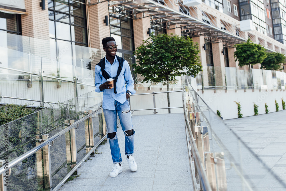 Photo of young American man in blue shirt holding coffee while walking in the street. Model. Outfit. Outside.