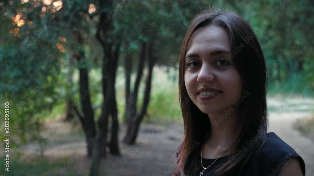 Portrait of cute positive young brunette girl smiling to the camera in the park with green trees in the evening, Slow motion Portrait of cute positive young brunette girl smiling to the camera in the park with green trees in the evening, Slow motion