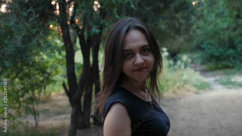 Attractive young brunette girl turning to left and right shoulder and smiling to the camera in the park with green trees in the evening, Slow motion Attractive young brunette girl turning to left and right shoulder and smiling to the camera in the park with green trees in the evening, Slow motion