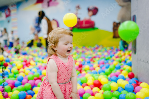 Portrait little cute baby girl princess infant 1-2 year standing and play with balloons, colorful balls in playground, ball pit, dry pool for birthday party. Celebration first year concept.
