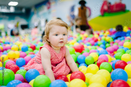 Happy laughing girl playing with toys, colorful balls in playground, ball pit, dry pool. Little cute child having fun in ball pit on birthday party in kids amusement park and play center indoor.