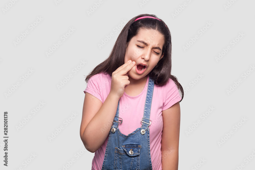 Tooth ache. Portrait of sick brunette young girl in pink t-shirt and blue overalls standing and touching her cheek because toothache. indoor studio shot, isolated on light gray background. Tooth ache. Portrait of sick brunette young girl in pink t-shirt and blue overalls standing and touching her cheek because toothache. indoor studio shot, isolated on light gray background.