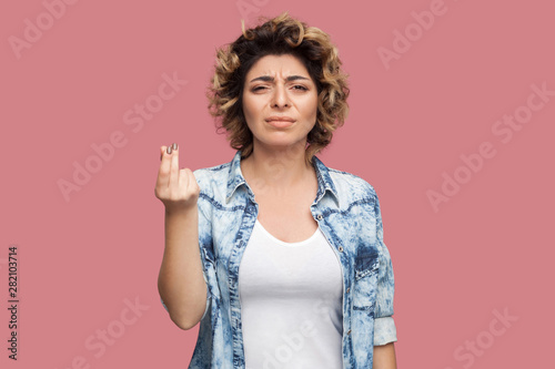 Photos Portrait of serious young woman with curly hairstyle in casual blue shirt standing with money or italian gesture and looking at camera