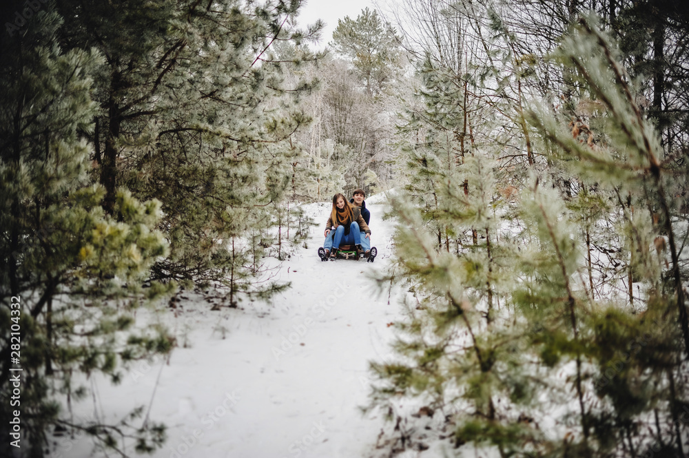 Obraz premium Smiling and happy couple enjoy in sledding on downhill in a forest or city park. Two young people sliding on a wooden sled at snow winter day.