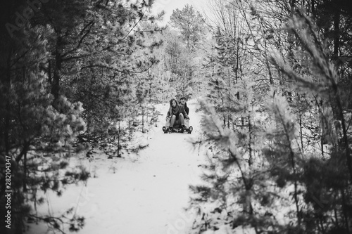 Smiling and happy couple enjoy in sledding on downhill in a forest or city park. Two young people: man with girl sliding on a wooden sled at snow winter day. Black and white photo.