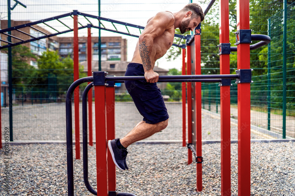 Strong muscular man doing push-ups on uneven bars in outdoor street gym. Workout lifestyle concept.