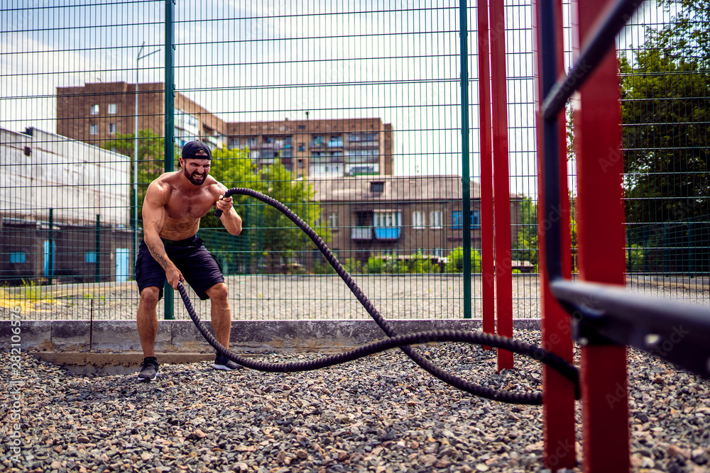 Fototapeta premium Athletic looking man working out with rope at street gym yard. Strength and motivation. Outdoor workout.