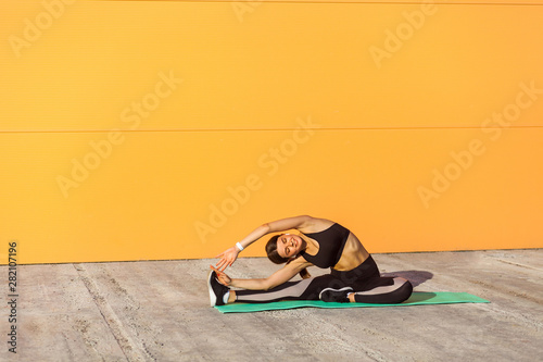 Young woman practicing yoga, doing revolved head to knee forward exercise, parivrtta janu sirsasana pose, working out, wearing sportswear, black pants and top, Outdoor, orange wall background,