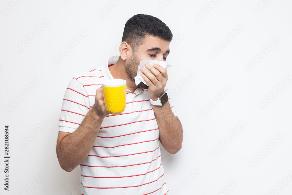 Cold and flu. Portrait of sick bearded young man in striped t-shirt standing, holding tissue, cleaning his nose, holding hot drink, try to be treated. indoor studio shot, isolated on white background. Cold and flu. Portrait of sick bearded young man in striped t-shirt standing, holding tissue, cleaning his nose, holding hot drink, try to be treated. indoor studio shot, isolated on white background.
