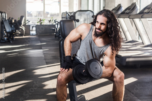 Portrait of confident young adult man with long curly hair athlete working out in gym, sitting on a bench and holding one dumbbell with raised arm, doing exercises for biceps. indoor, looking away