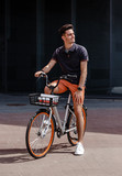 Stylish guy dressed in trendy casual clothes rides bicycle in the street against the background of a gray building