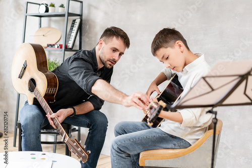Adult man explains his son, who learn to play guitar, how to play studied chord correctly. Education with family