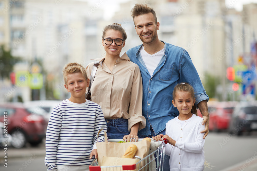 Obraz premium Portrait of contemporary family with two kids posing with shopping cart at parking lot outdoors, copy space