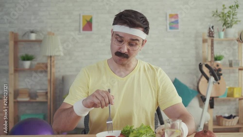 Happy and joyful young sporty man with a mustache eating vegetables sitting at the table