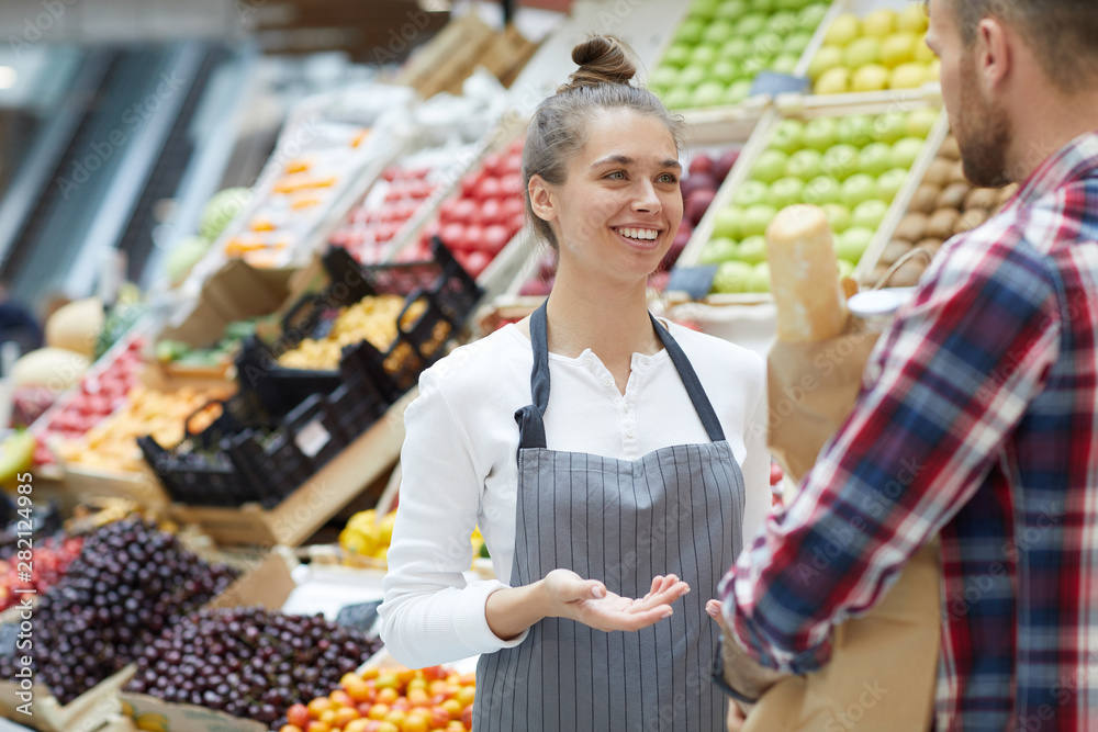 Waist up portrait of pretty young saleswoman talking to customer while standing by fresh fruits and vegetables at farmers market, copy space Waist up portrait of pretty young saleswoman talking to customer while standing by fresh fruits and vegetables at farmers market, copy space