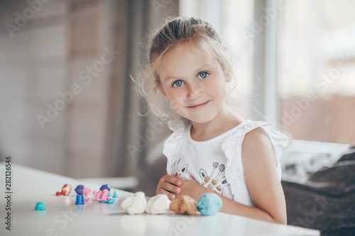 Little girl is playing with plasticine while sitting at table against the background of a large window