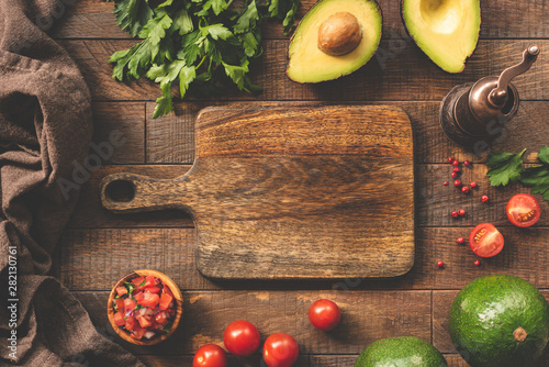 Fresh vegetables and cutting board for cooking, food frame background. Avocado, tomato, salsa, parsley and pepper spices on wooden table