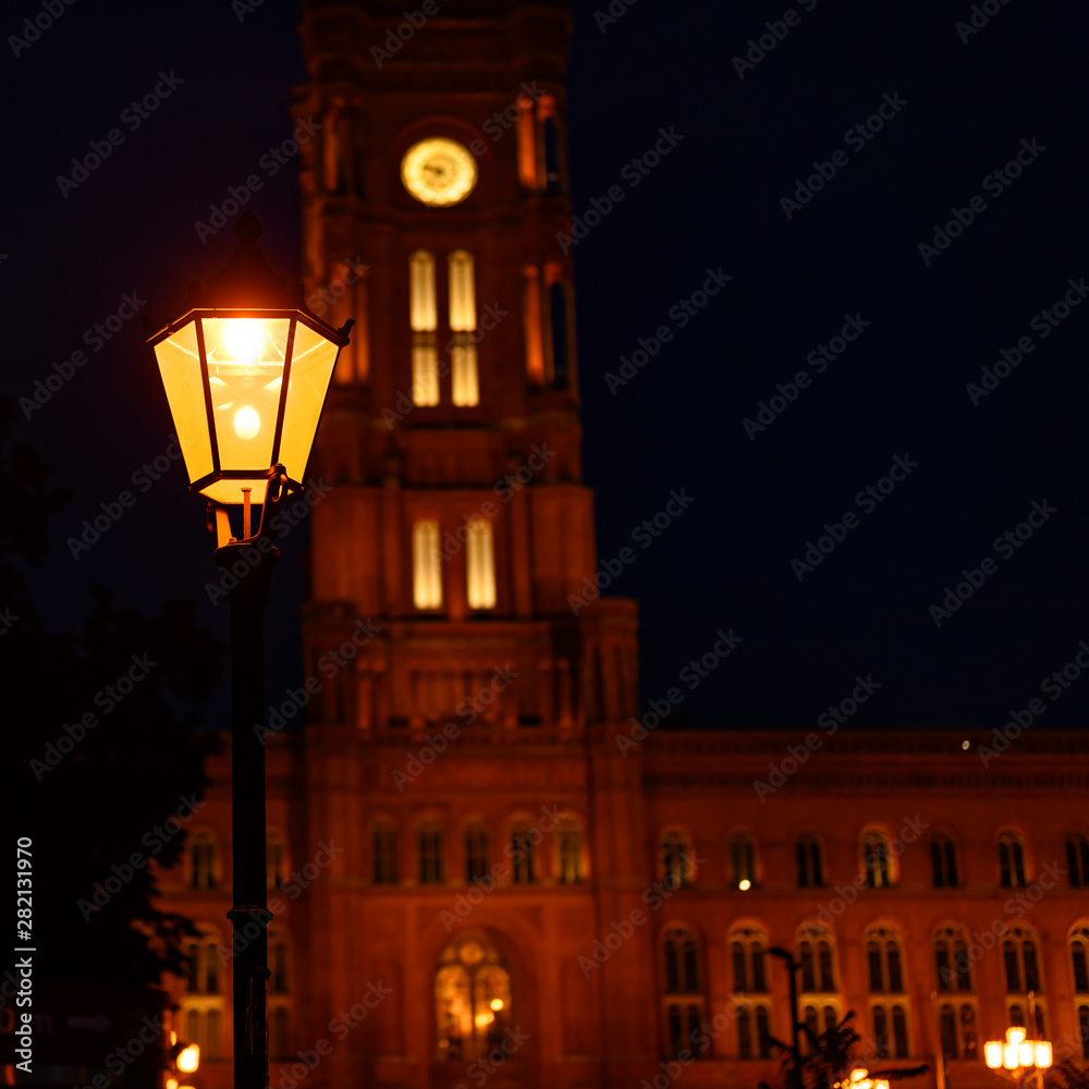 The Red Town Hall in the center of Berlin at night. In the foreground is a historic Berlin lantern on which the focus lies. The town hall is out of focus in the background.