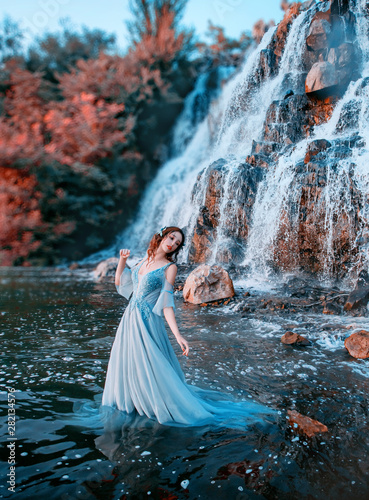 Fototapeta Naklejka Na Ścianę i Meble -  lovely girl with fair tender skin stands in aqua in long light blue dress, mystical mermaid near high waterfall with powerful streams of cold water, Lost doll with Korean face and waterproof makeup