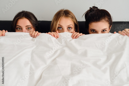 Three young beautiful women sitting in bed and covering faces with white blanket dreamily looking in camera together in modern cozy hotel