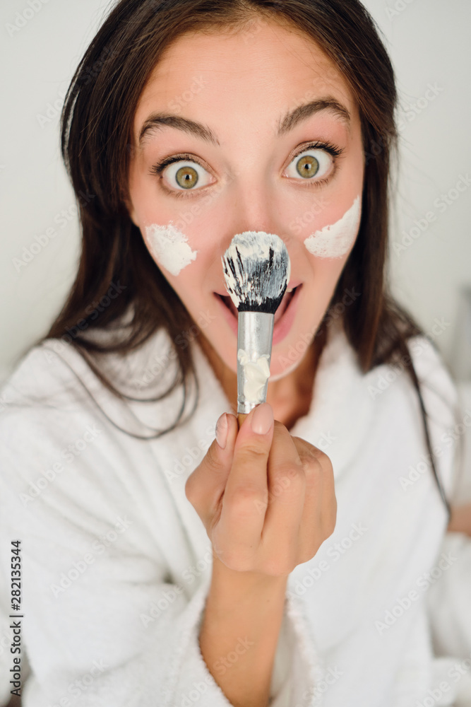 Young pretty joyful woman in white bathrobe with cosmetic mask on face holding brush near nose happily looking in camera in modern hotel Young pretty joyful woman in white bathrobe with cosmetic mask on face holding brush near nose happily looking in camera in modern hotel