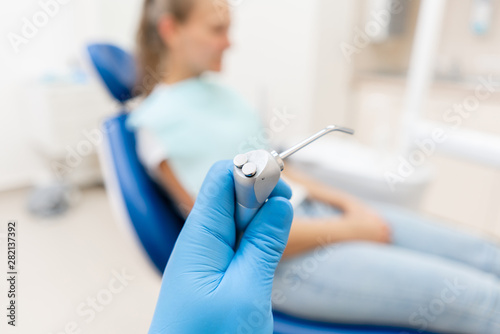 Close-up hand of dentist in the glove holds Gun dental water and air. The patient in blue chair at the background. Office where dentist conducts inspection and concludes.