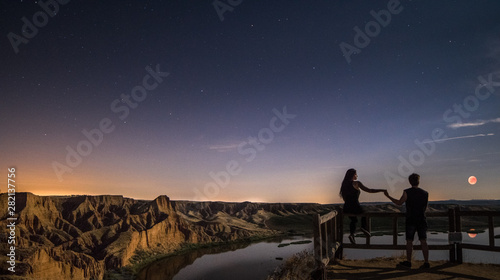 A couple looking the lunar eclipse in a canyon. River crossing below them.