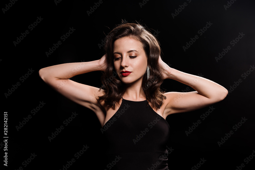 young woman in dress standing and touching hair isolated on black