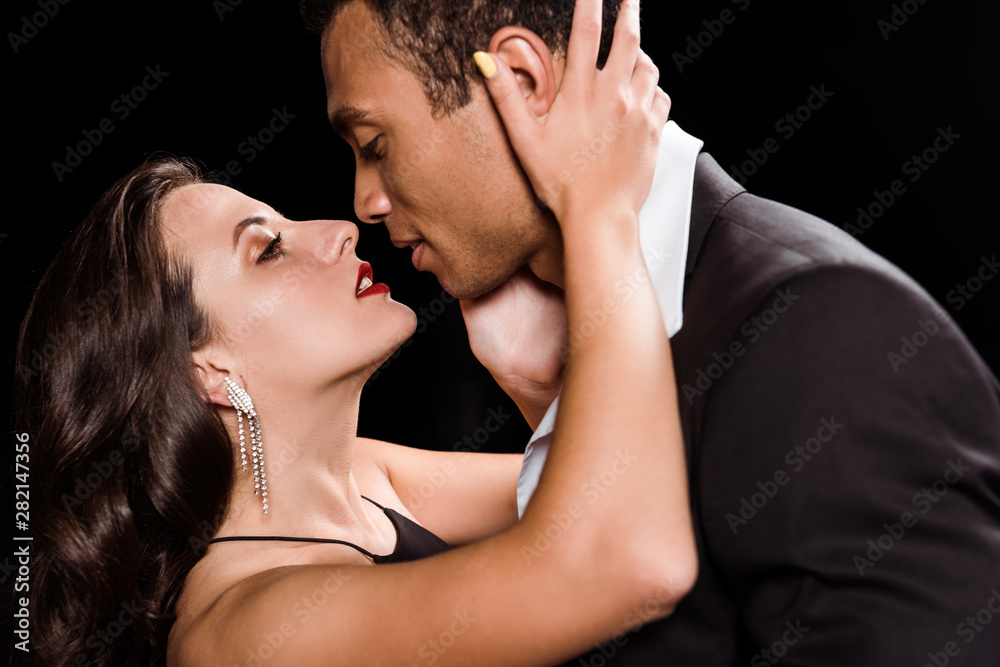 young woman hugging and looking at handsome mixed race man isolated on black young woman hugging and looking at handsome mixed race man isolated on black