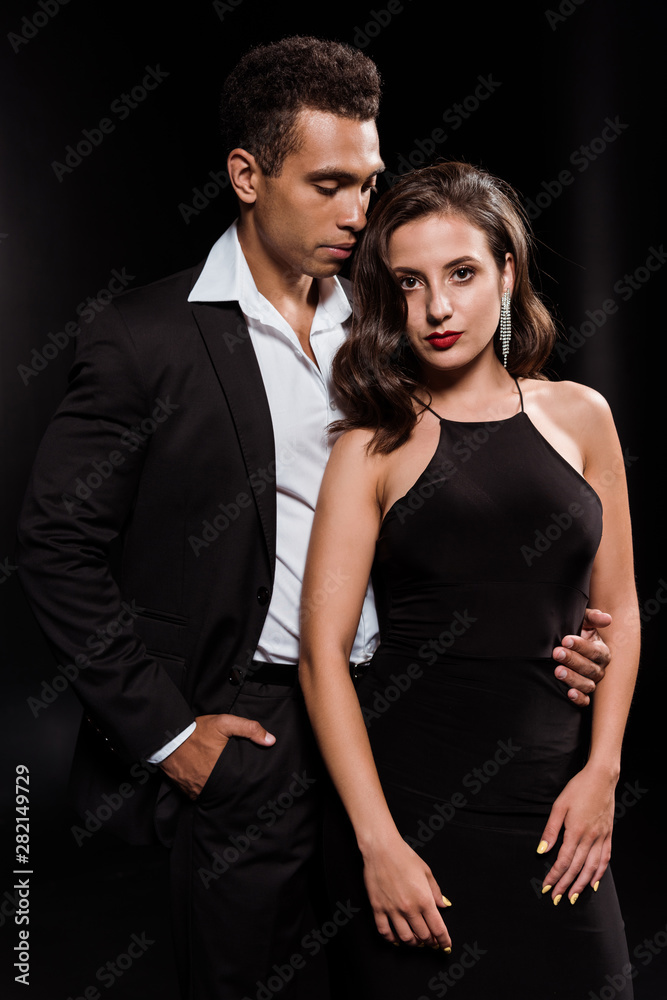 handsome mixed race man standing with hand in pocket and looking at attractive woman isolated on black handsome mixed race man standing with hand in pocket and looking at attractive woman isolated on black