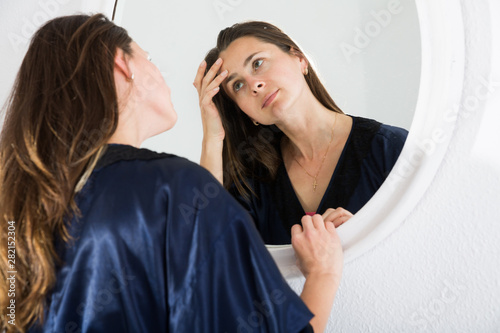 Young female touching face and looking at mirror in bedroom