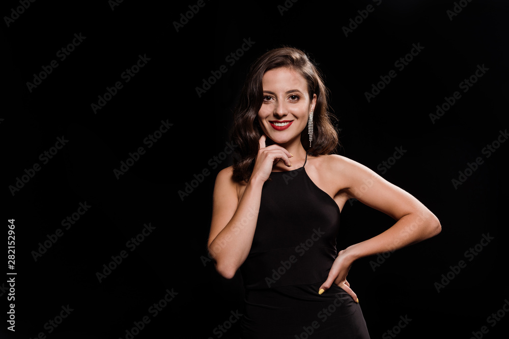 happy woman in dress standing with hand on hip isolated on black