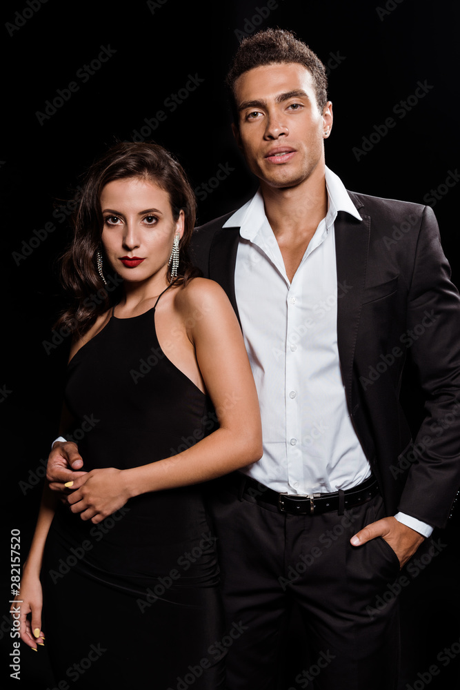 handsome mixed race man standing with hand in pocket near attractive woman isolated on black handsome mixed race man standing with hand in pocket near attractive woman isolated on black