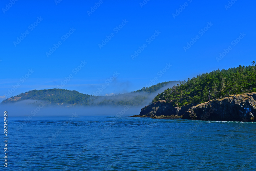 Fototapeta premium Morning fog hugs the coast of Deception Pass as it enters the Salish Sea in Washington State