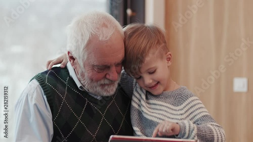 Smiling grandfather and grandson using digital tablet for surfing internet and playing game near the fireplace at home grandpa adult grandchild child childhood communication computer photo