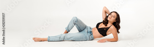 panoramic shot of attractive woman with barefoot lying in blue jeans and touching hair on white