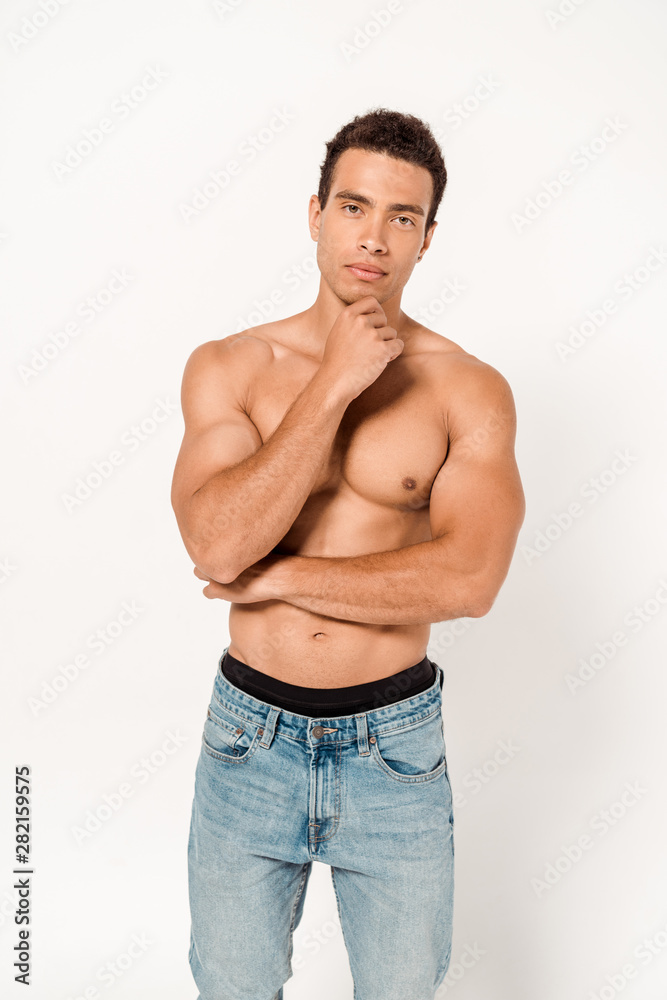 pensive mixed race man touching chin while standing in blue jeans on white