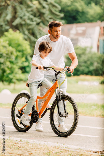 full length view of father looking forward and helping son to ride on bike while son sitting on bicycle