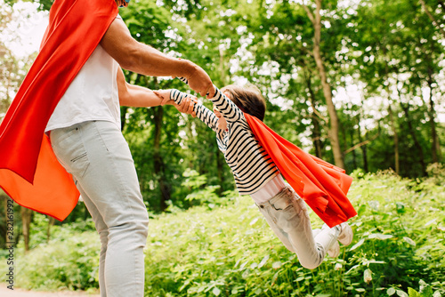 cropped view of father spinning little boy in red superhero cloak