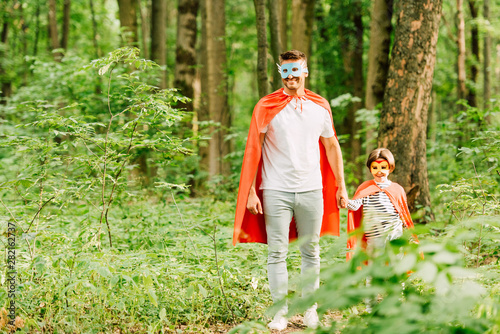 full length view of father and son in superhero costumes holding hands while standing in green forest