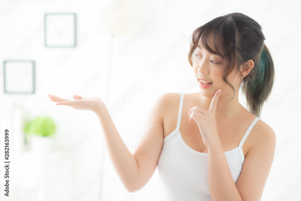 Beautiful young asian woman smiling and showing skin care product for advertisement at bedroom, beauty asia girl presenting makeup cosmetic for advertise with surprise on palms at room. Beautiful young asian woman smiling and showing skin care product for advertisement at bedroom, beauty asia girl presenting makeup cosmetic for advertise with surprise on palms at room.