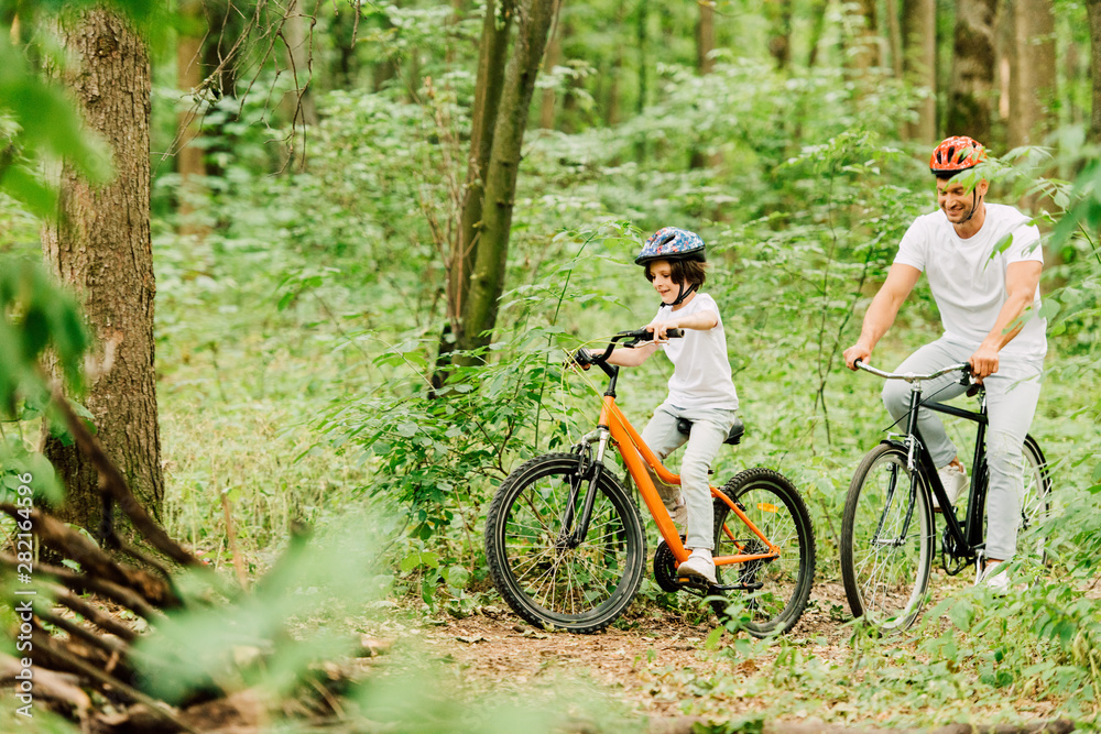Selective focus of father and son in helmets riding bicycles around forest